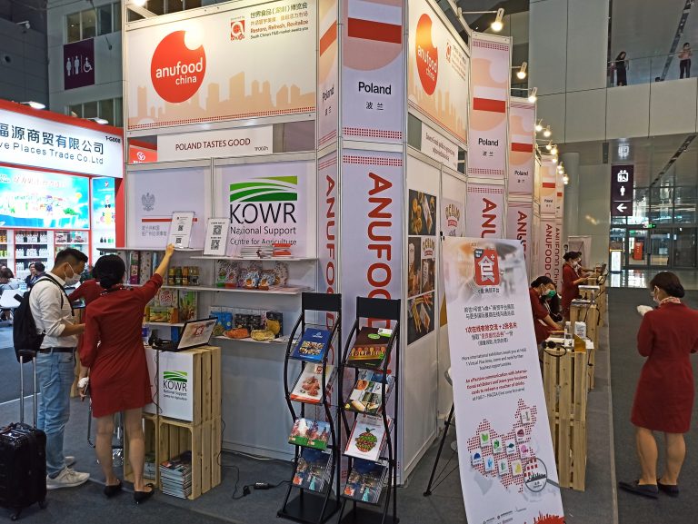 Zdjęcie przedstawia polskie stoisko narodowe na targach Anufood 2021. Na stoisku dominują barwy biała i czerwona. Na scianach stoiska są umieszczone logotypy i grafiki firm uczestniczących w stoisku. Przedstawiciele firm siedzą przy ladach informacyjnych znajdujących się przy krawędzi stoiska, promując swoje produkty i prowadzą rozmowy ze zwiedzającymi targi. Na stoisku Znajduje się również punkt informacyjny o działaniach prowadzonych przez Krajowy Ośrodek Wsparcia Rolnictwa pod hasłem Poland tastes good.