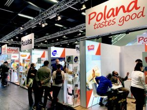 Zdjęcie przedstawia polskie stoisko narodowe. Na stoisku przeważają kolory biały i czerwony. Na fryzie stoiska dobrze widoczne logotypy Polska i Poland tastes good. Na stoisku przedstawiciele polskich firm promują swoje produkty. Stoisko cieszy się dużym zainteresowaniem zwiedzających.